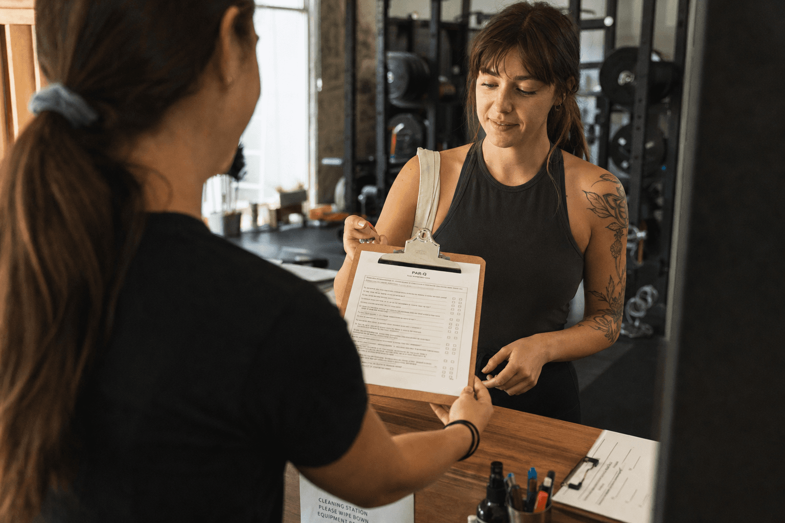 A trainer handing a new client a PAR-Q questionnaire on a clipboard at the gym
